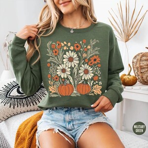 K&ouml;nnte beinhalten: Olivgr&uuml;nes Sweatshirt mit Blumen- und K&uuml;rbisdesign. Die Grafik zeigt gro&szlig;e wei&szlig;e G&auml;nsebl&uuml;mchen, orangefarbene Blumen und zwei orangefarbene K&uuml;rbisse. Das Sweatshirt hat lange &Auml;rmel und einen Rundhalsausschnitt. Das Wort "MILITARY GREEN" ist auf einem kleinen Etikett aufgedruckt.