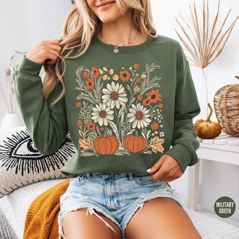 K&ouml;nnte beinhalten: Olivgr&uuml;nes Sweatshirt mit Blumen- und K&uuml;rbisdesign. Die Grafik zeigt gro&szlig;e wei&szlig;e G&auml;nsebl&uuml;mchen, orangefarbene Blumen und zwei orangefarbene K&uuml;rbisse. Das Sweatshirt hat lange &Auml;rmel und einen Rundhalsausschnitt. Das Wort "MILITARY GREEN" ist auf einem kleinen Etikett aufgedruckt.