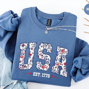 May include: Indigo blue crewneck sweatshirt with the word "USA" in a floral pattern. The sweatshirt also features the text "EST. 1776" in red. A silver necklace with a pendant is draped across the sweatshirt.