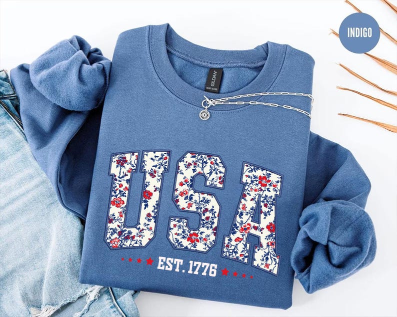 May include: Indigo blue crewneck sweatshirt with the word "USA" in a floral pattern. The sweatshirt also features the text "EST. 1776" in red. A silver necklace with a pendant is draped across the sweatshirt.