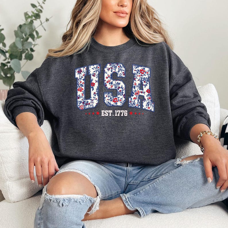 May include: Dark gray sweatshirt with the word "USA" in a floral design, featuring red, white, and blue flowers. The text "EST. 1776" is below the word "USA". The sweatshirt is a crew neck style.