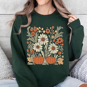 K&ouml;nnte beinhalten: Ein dunkelgr&uuml;nes Sweatshirt mit einem Blumendesign mit G&auml;nsebl&uuml;mchen, orangefarbenen Blumen und K&uuml;rbissen. Das Design ist auf der Vorderseite zentriert. Das Sweatshirt hat einen Rundhalsausschnitt.