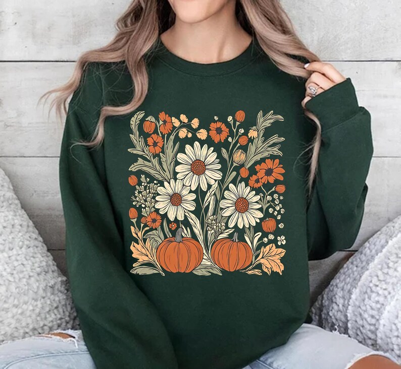 K&ouml;nnte beinhalten: Ein dunkelgr&uuml;nes Sweatshirt mit einem Blumendesign mit G&auml;nsebl&uuml;mchen, orangefarbenen Blumen und K&uuml;rbissen. Das Design ist auf der Vorderseite zentriert. Das Sweatshirt hat einen Rundhalsausschnitt.