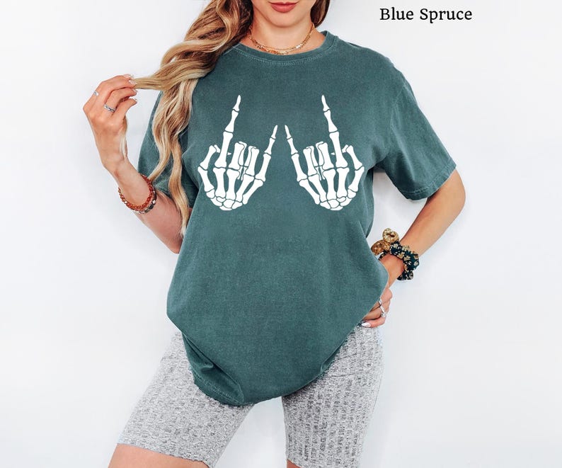 Puede incluir: Una camiseta de manga corta de color azul abeto con un estampado gr&aacute;fico blanco de dos manos de esqueleto haciendo un gesto de rock and roll. La camiseta tiene cuello redondo y un corte relajado, con el texto Blue Spruce impreso en la esquina.