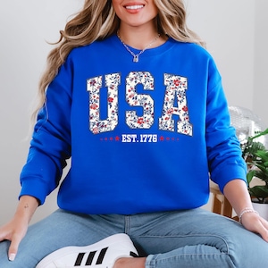 May include: A royal blue sweatshirt with the word "USA" in a floral design. The letters are filled with a white background and red and blue flowers. Below the word "USA" is the text "EST. 1776" with red stars.