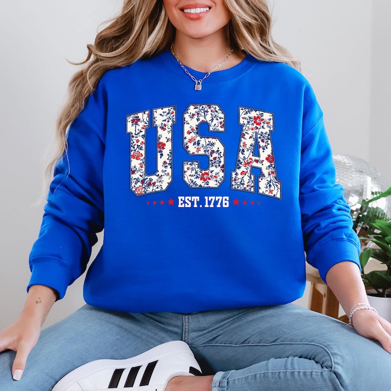 May include: A royal blue sweatshirt with the word "USA" in a floral design. The letters are filled with a white background and red and blue flowers. Below the word "USA" is the text "EST. 1776" with red stars.