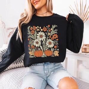 K&ouml;nnte beinhalten: Schwarzes Langarm-Sweatshirt mit Blumen- und K&uuml;rbisdesign. Die Grafik zeigt wei&szlig;e G&auml;nsebl&uuml;mchen, orangefarbene Blumen und zwei orangefarbene K&uuml;rbisse. Das Design ist vor einem schwarzen Hintergrund gehalten und schafft eine herbstliche &Auml;sthetik.