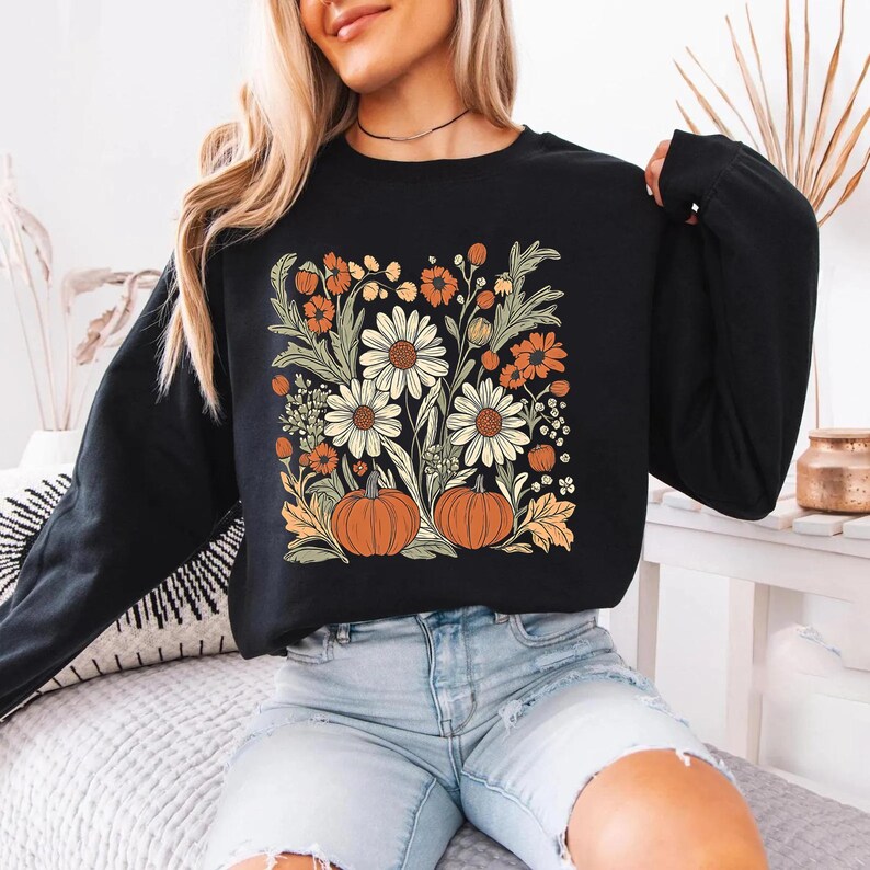 K&ouml;nnte beinhalten: Schwarzes Langarm-Sweatshirt mit Blumen- und K&uuml;rbisdesign. Die Grafik zeigt wei&szlig;e G&auml;nsebl&uuml;mchen, orangefarbene Blumen und zwei orangefarbene K&uuml;rbisse. Das Design ist vor einem schwarzen Hintergrund gehalten und schafft eine herbstliche &Auml;sthetik.