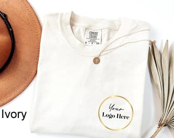 May include: Ivory-colored Comfort Colors t-shirt with a gold necklace and a gold-framed logo that says "Your Logo Here." A brown hat and dried plant stems are also visible.