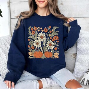 K&ouml;nnte beinhalten: Marineblaues Sweatshirt mit einem Blumendesign mit G&auml;nsebl&uuml;mchen, orangefarbenen Blumen und K&uuml;rbissen. Das Design ist mittig auf der Vorderseite. Das Sweatshirt ist aus weichem Material.