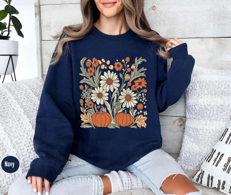 K&ouml;nnte beinhalten: Marineblaues Sweatshirt mit einem Blumendesign mit G&auml;nsebl&uuml;mchen, orangefarbenen Blumen und K&uuml;rbissen. Das Design ist mittig auf der Vorderseite. Das Sweatshirt ist aus weichem Material.