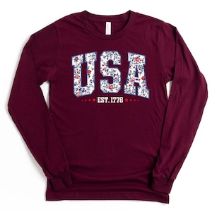 May include: A long-sleeve maroon shirt with the word "USA" in a floral design. The text "EST. 1776" is below the word "USA". The shirt has a crew neck and long sleeves.