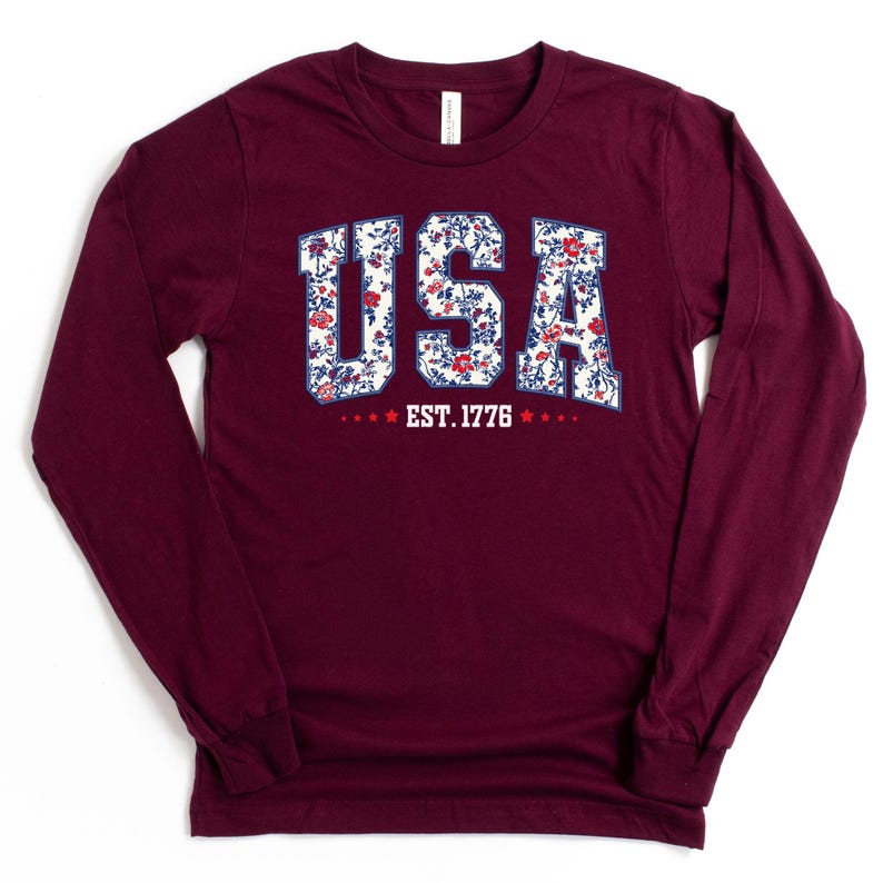 May include: A long-sleeve maroon shirt with the word "USA" in a floral design. The text "EST. 1776" is below the word "USA". The shirt has a crew neck and long sleeves.