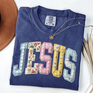 May include: A blue short-sleeved t-shirt featuring the word JESUS in large, fabric-appliqued letters. Each letter has a unique pattern, including blue gingham, floral print, pink polka dots, yellow daisies, and light blue patchwork fabric.
