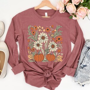 K&ouml;nnte beinhalten: Ein lang&auml;rmeliges, malvenfarbenes T-Shirt mit einem Blumendesign mit G&auml;nsebl&uuml;mchen, orangefarbenen Blumen und K&uuml;rbissen. Das Design ist auf dem Shirt zentriert, mit hochgekrempelten &Auml;rmeln. Das Shirt wird auf einem Holzb&uuml;gel pr&auml;sentiert.