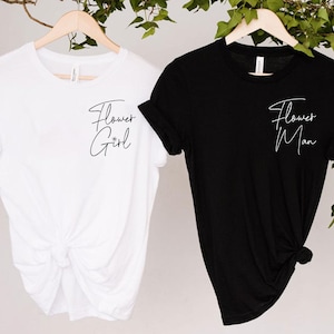 May include: Two t-shirts, one white and one black, hanging on wooden hangers. The white shirt has the text "Flower Girl" in cursive, and the black shirt has "Flower Man". Both shirts are short-sleeved.
