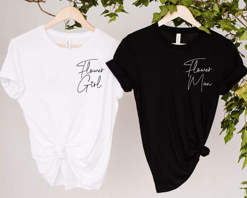 May include: Two t-shirts, one white and one black, hanging on wooden hangers. The white shirt has the text "Flower Girl" in cursive, and the black shirt has "Flower Man". Both shirts are short-sleeved.