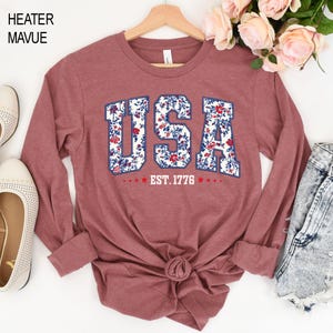 May include: A long-sleeved, heather mauve shirt with the word "USA" in a floral design. The text "EST. 1776" is below the word "USA". The shirt is displayed on a wooden hanger.