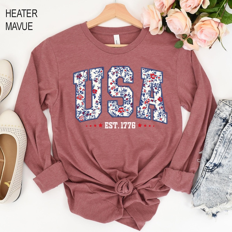 May include: A long-sleeved, heather mauve shirt with the word "USA" in a floral design. The text "EST. 1776" is below the word "USA". The shirt is displayed on a wooden hanger.