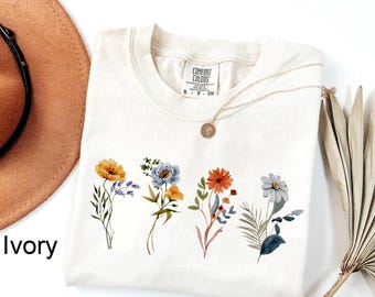 May include: Ivory-colored t-shirt with a crew neck, featuring a floral design of watercolor flowers in yellow, blue, orange, and gray. A gold necklace with a pendant rests on the shirt. The shirt is labeled "Comfort Colors".