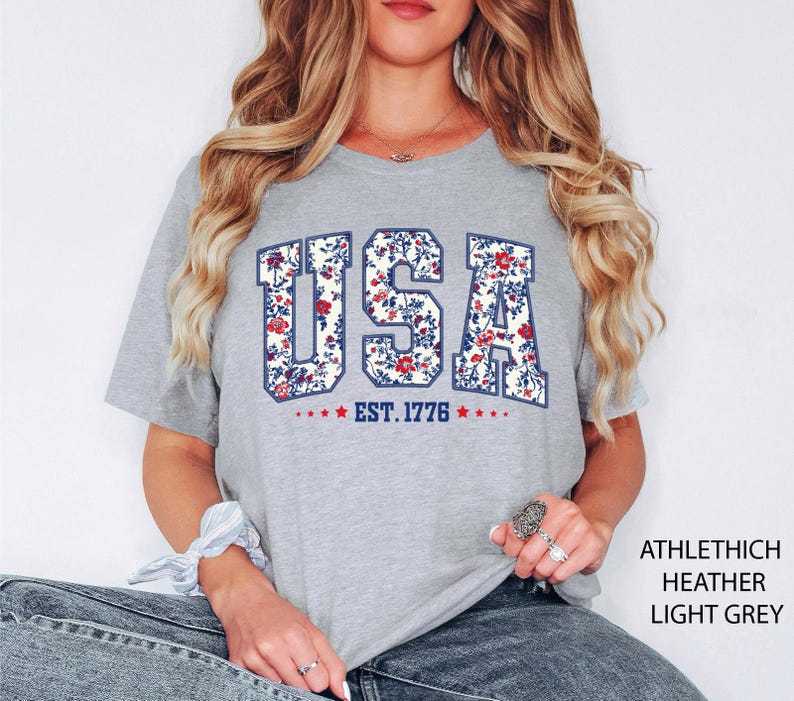 May include: Light grey t-shirt with the word "USA" in a floral design, with the text "EST. 1776" below. The shirt is made of a soft material and is a heather light grey color. The shirt is a casual style.