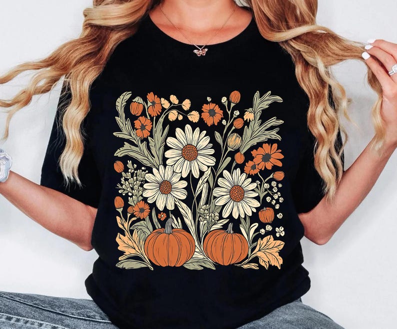 May include: Black t-shirt featuring a floral design with daisies, orange flowers, and pumpkins. The design includes green leaves and stems, creating a fall or autumn theme. The shirt is a classic crew neck style.