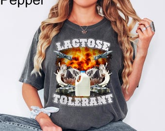May include: Dark gray t-shirt with a graphic design. The design features the words "LACTOSE TOLERANT" above and below a central image. The image includes cows, a milk jug, jets, and an explosion.