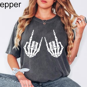 Puede incluir: Una camiseta de manga corta gris carb&oacute;n con un estampado gr&aacute;fico blanco de dos manos de esqueleto haciendo un gesto de rock and roll. La camiseta tiene un acabado lavado de estilo vintage y la palabra Pepper est&aacute; impresa en la esquina superior izquierda.