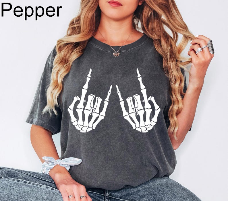 Puede incluir: Una camiseta de manga corta gris carb&oacute;n con un estampado gr&aacute;fico blanco de dos manos de esqueleto haciendo un gesto de rock and roll. La camiseta tiene un acabado lavado de estilo vintage y la palabra Pepper est&aacute; impresa en la esquina superior izquierda.