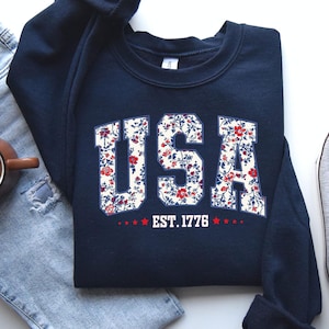 May include: Navy blue sweatshirt with the word "USA" in white block letters filled with a floral pattern of red and blue flowers. Below the text is "EST. 1776" with red stars. The sweatshirt is folded.