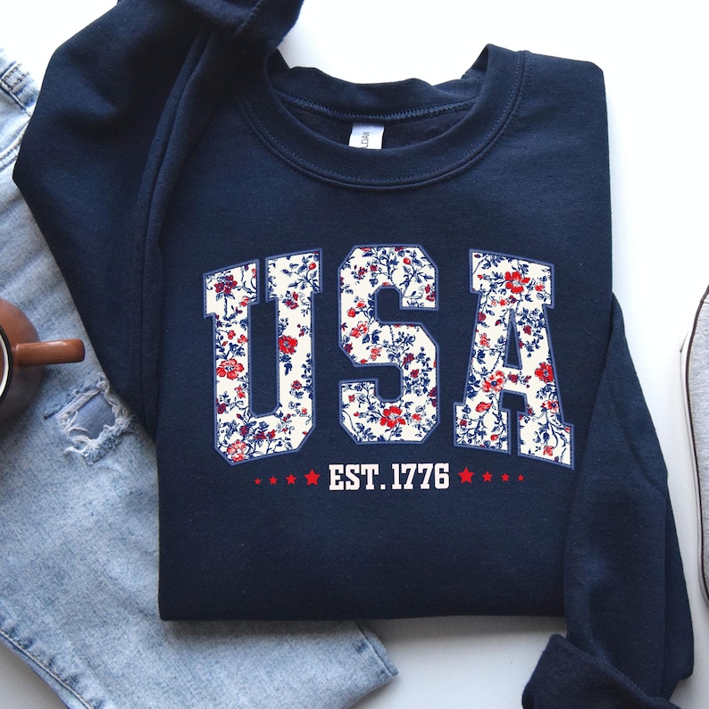 May include: Navy blue sweatshirt with the word "USA" in white block letters filled with a floral pattern of red and blue flowers. Below the text is "EST. 1776" with red stars. The sweatshirt is folded.