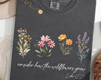 May include: A dark gray t-shirt with a floral design and the text "consider how the wildflowers grow Luke 12:27." The shirt features a crew neck and is adorned with a gold necklace. The word "Pepper" is printed at the bottom.