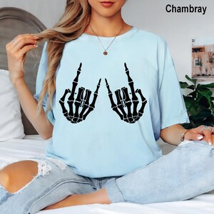 Puede incluir: Una camiseta azul claro con un estampado gr&aacute;fico negro de dos manos de esqueleto haciendo el signo de mano de rock and roll. La camiseta tiene cuello redondo y mangas cortas. El texto Chambray est&aacute; impreso en la esquina superior derecha.