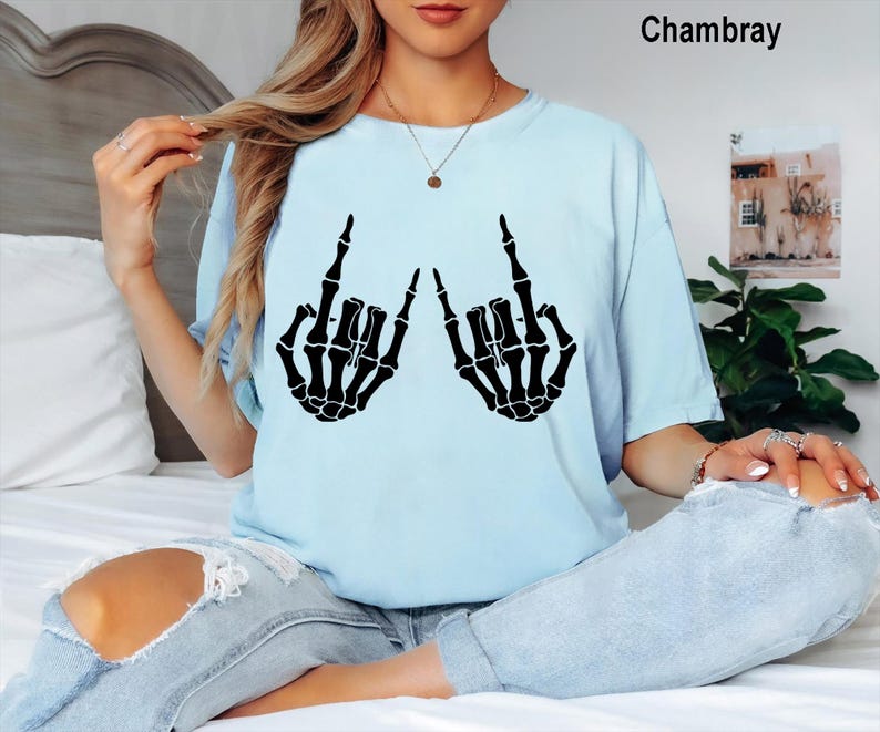 Puede incluir: Una camiseta azul claro con un estampado gr&aacute;fico negro de dos manos de esqueleto haciendo el signo de mano de rock and roll. La camiseta tiene cuello redondo y mangas cortas. El texto Chambray est&aacute; impreso en la esquina superior derecha.
