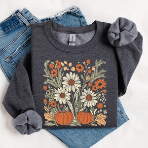 K&ouml;nnte beinhalten: Dunkelgraues Sweatshirt mit einem Blumendesign mit wei&szlig;en G&auml;nsebl&uuml;mchen, orangefarbenen Blumen und zwei K&uuml;rbissen. Das Design ist auf der Brust zentriert. Das Sweatshirt ist auf einer blauen Jeans gefaltet.