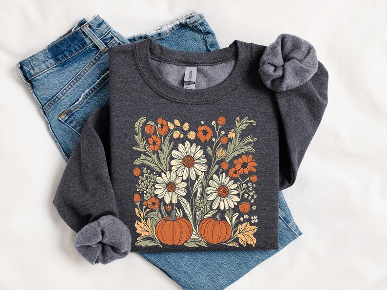 K&ouml;nnte beinhalten: Dunkelgraues Sweatshirt mit einem Blumendesign mit wei&szlig;en G&auml;nsebl&uuml;mchen, orangefarbenen Blumen und zwei K&uuml;rbissen. Das Design ist auf der Brust zentriert. Das Sweatshirt ist auf einer blauen Jeans gefaltet.