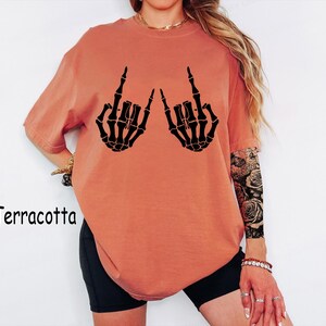 Puede incluir: Una camiseta de manga corta color terracota con un estampado gr&aacute;fico negro de dos manos esquel&eacute;ticas haciendo el gesto de rock and roll. La camiseta tiene cuello redondo y un corte holgado. La palabra Terracotta est&aacute; impresa a la izquierda.
