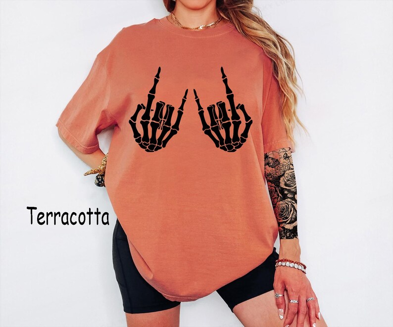 Puede incluir: Una camiseta de manga corta color terracota con un estampado gr&aacute;fico negro de dos manos esquel&eacute;ticas haciendo el gesto de rock and roll. La camiseta tiene cuello redondo y un corte holgado. La palabra Terracotta est&aacute; impresa a la izquierda.