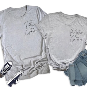 May include: Two gray t-shirts with the phrases "Father of the Groom" and "Mother of the Groom" in black script. A pair of black and white sneakers and a pair of blue denim shorts are also in the image.