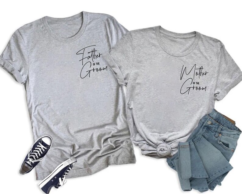 May include: Two gray t-shirts with the phrases "Father of the Groom" and "Mother of the Groom" in black script. A pair of black and white sneakers and a pair of blue denim shorts are also in the image.