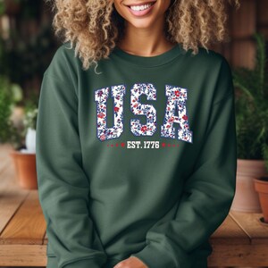 May include: Dark green sweatshirt with the word "USA" in white block letters filled with a floral pattern of red and blue flowers. Below the word "USA" is the text "EST. 1776" in white.