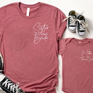 May include: A dusty rose t-shirt with the text "Sister of the Bride" and a matching baby onesie with "Mother of the Flower Girl" printed on it. Black and white sneakers are also in the image.