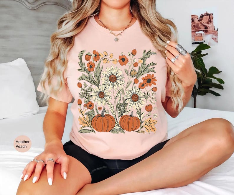 K&ouml;nnte beinhalten: Pfirsichfarbenes T-Shirt mit floralem Design mit K&uuml;rbissen, G&auml;nsebl&uuml;mchen und anderen Blumen in Orange, Gelb und Gr&uuml;n. Das Shirt hat kurze &Auml;rmel und einen Rundhalsausschnitt. Der Text "Heather Peach" befindet sich auf der linken Seite des Bildes.