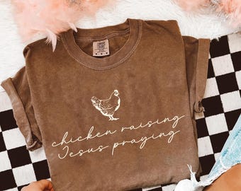 May include: A brown short-sleeve t-shirt featuring a white graphic of a chicken centered above the text "chicken raising Jesus praying" in a white cursive font. The shirt has a crew neckline and a Comfort Colors brand label at the inner neck.
