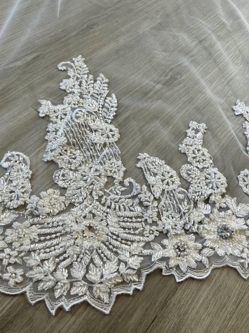 Op de afbeelding: Witte bruids sluier met een parelversierde kant rand. De kant heeft een bloemmotief met parel-achtige versieringen.