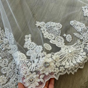 Op de afbeelding: Witte bruids sluier met delicate bloemen borduurwerk en parel versieringen. De sluier heeft een gegolfde rand en is gemaakt van zachte, transparante tule.