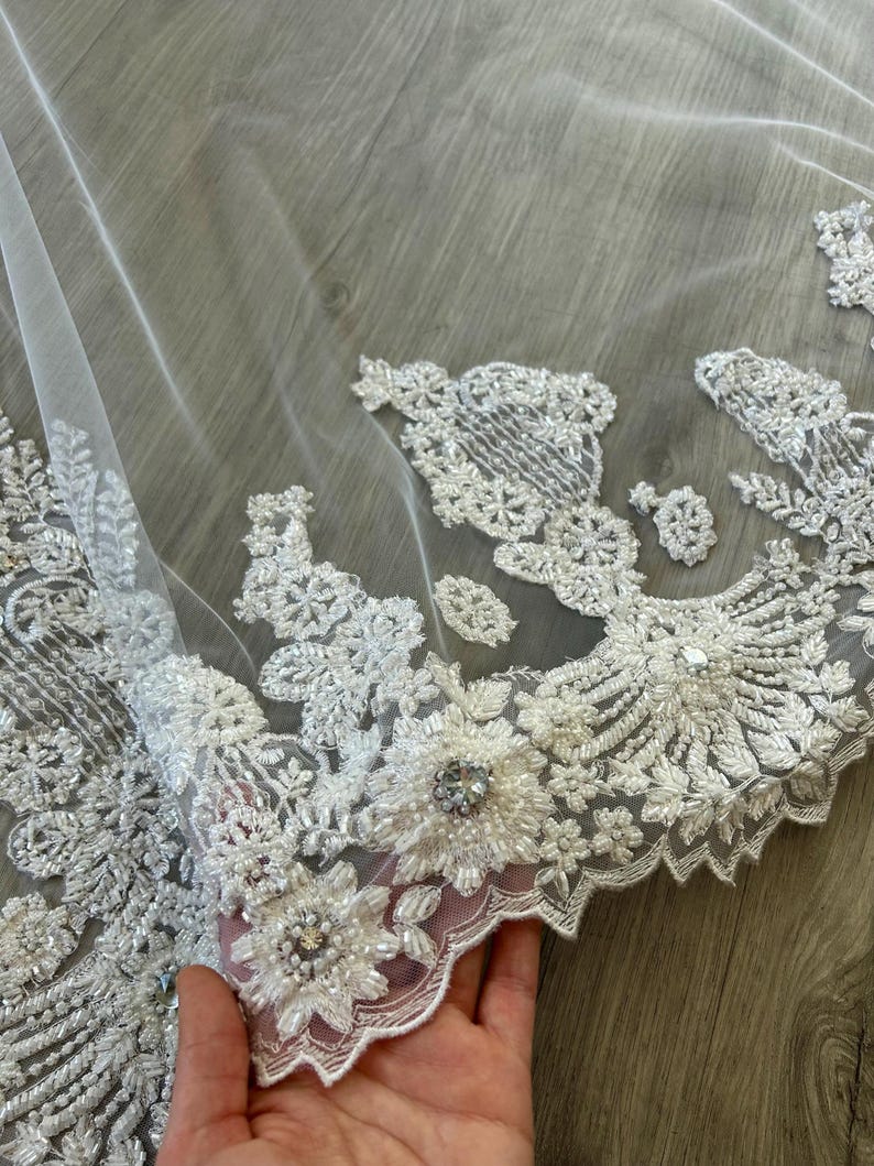 Op de afbeelding: Witte bruids sluier met delicate bloemen borduurwerk en parel versieringen. De sluier heeft een gegolfde rand en is gemaakt van zachte, transparante tule.