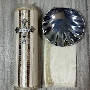May include: A baptism candle with a silver cross and decorative trim, next to a silver shell-shaped bowl and a cream-colored cloth with lace trim. The candle is beige with brown and cream stripes.