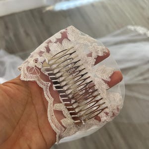 Op de afbeelding: Een witte bruids sluier met delicate kant en een zilveren kam eraan bevestigd. De sluier is gemaakt van zacht tule en heeft een delicaat bloemenpatroon.