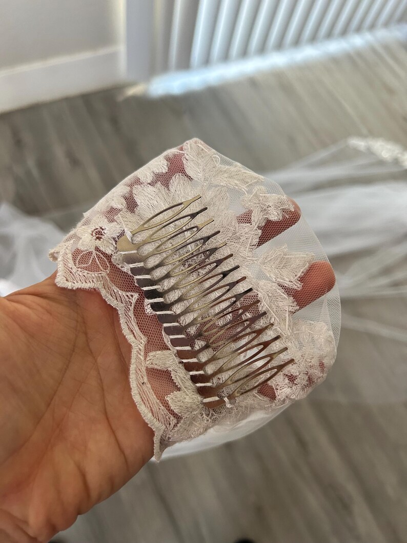 Op de afbeelding: Een witte bruids sluier met delicate kant en een zilveren kam eraan bevestigd. De sluier is gemaakt van zacht tule en heeft een delicaat bloemenpatroon.